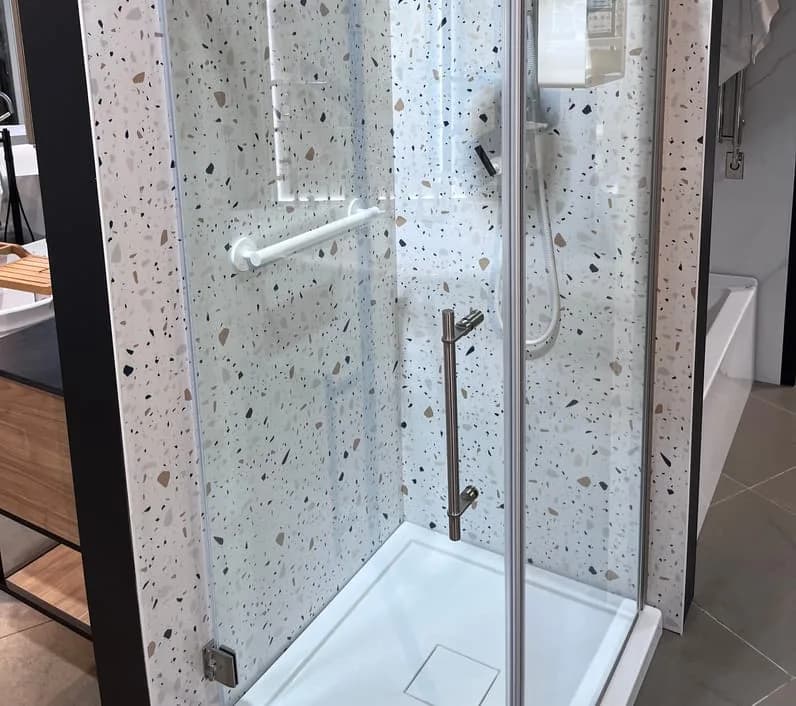 Douche vitrée sur base blanche, murs effet terrazzo et barre d’appui